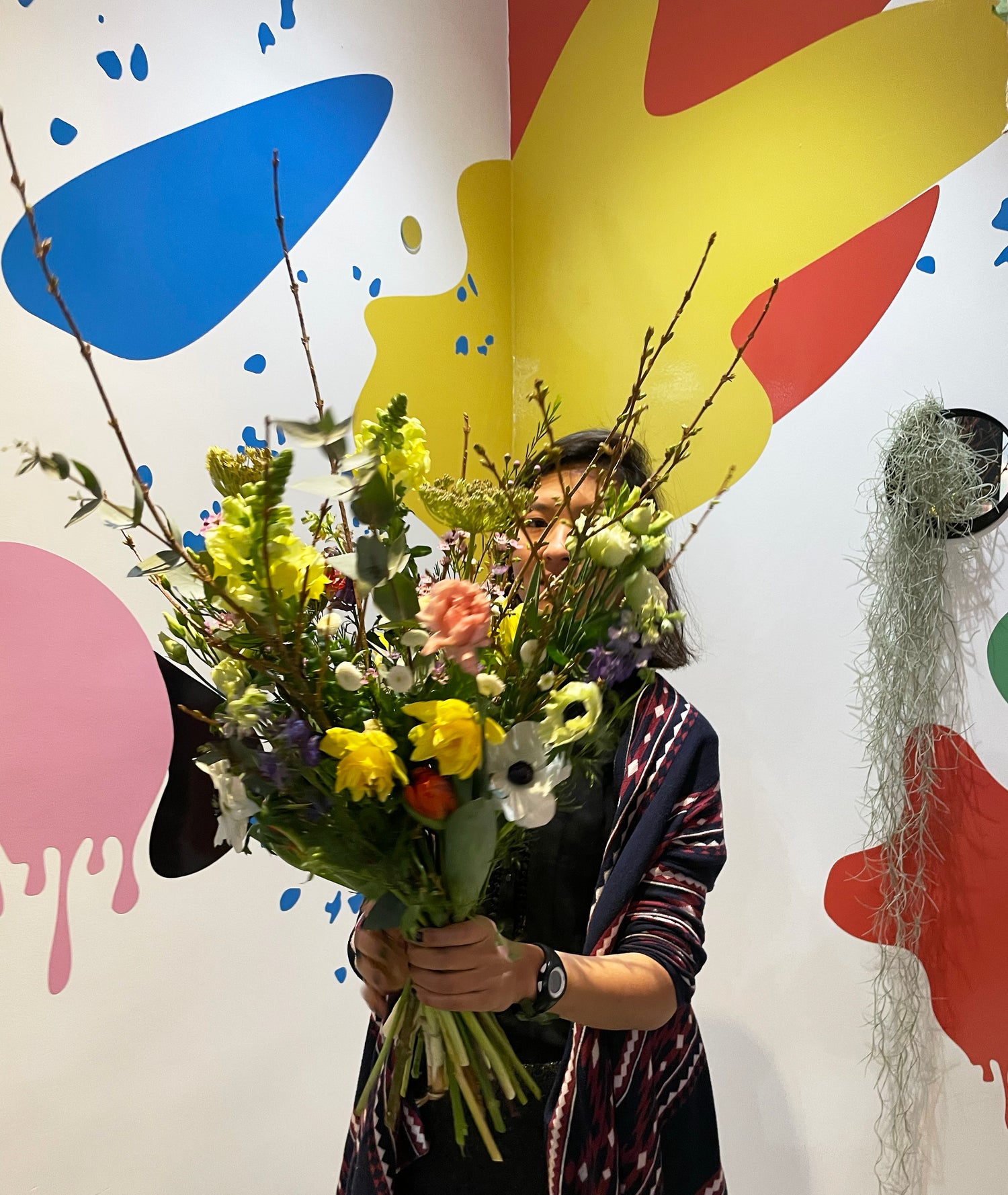 Une femme tient un gros bouquet de fleurs devant elle, le mur du fond est plein de couleurs